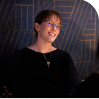 A woman, Tracey Schneider, with glasses smiles, wearing a black shirt, against a dark blue and gold patterned background.