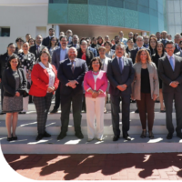 A group of people in formal attire stand together, casting long shadows on a tiled floor. Some individuals are wearing suits and blazers, while others are in dresses and skirts.