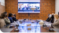 A group of people sit around a conference table, looking at a large screen displaying a video conference. The screen shows two main participants on the left and a wider view of a meeting room on the right.
