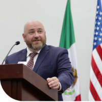 A man in a suit stands at a podium with the US and Mexican flags in the background.