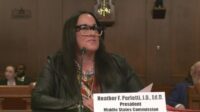 A speaker is speaking into a microphone. The nameplate in front of her reads “Heather F. Perfetti, J.D., Ed.D., President. Middle States Commission on Higher Education."